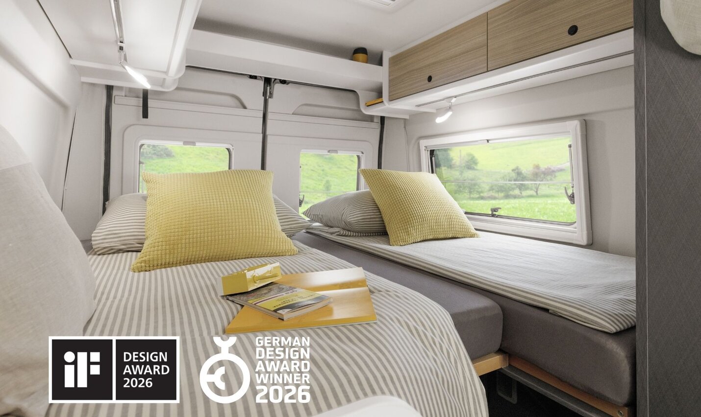 Sleeping area of a Campervan Fiat Plus with double bed, gray bedding, striped pillow, and view through window of green landscape.